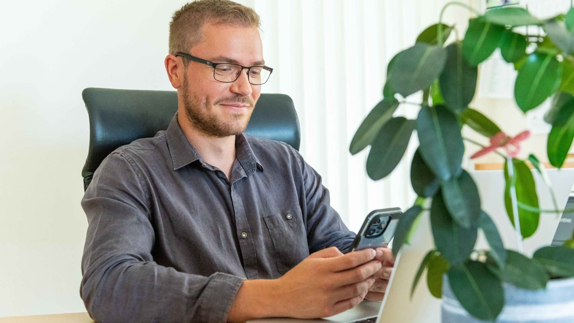 Mann schaut auf sein Handy, sitzt an einem Bürotisch mit Laptop und Blume, nutzt Timework von asut computer.