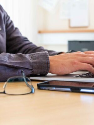 Männliche Hände arbeiten auf einem Notebook, Brille und Handy liegen auf dem Tisch, Timework-Software für Auftragszeiten und Projektzeiten geöffnet.